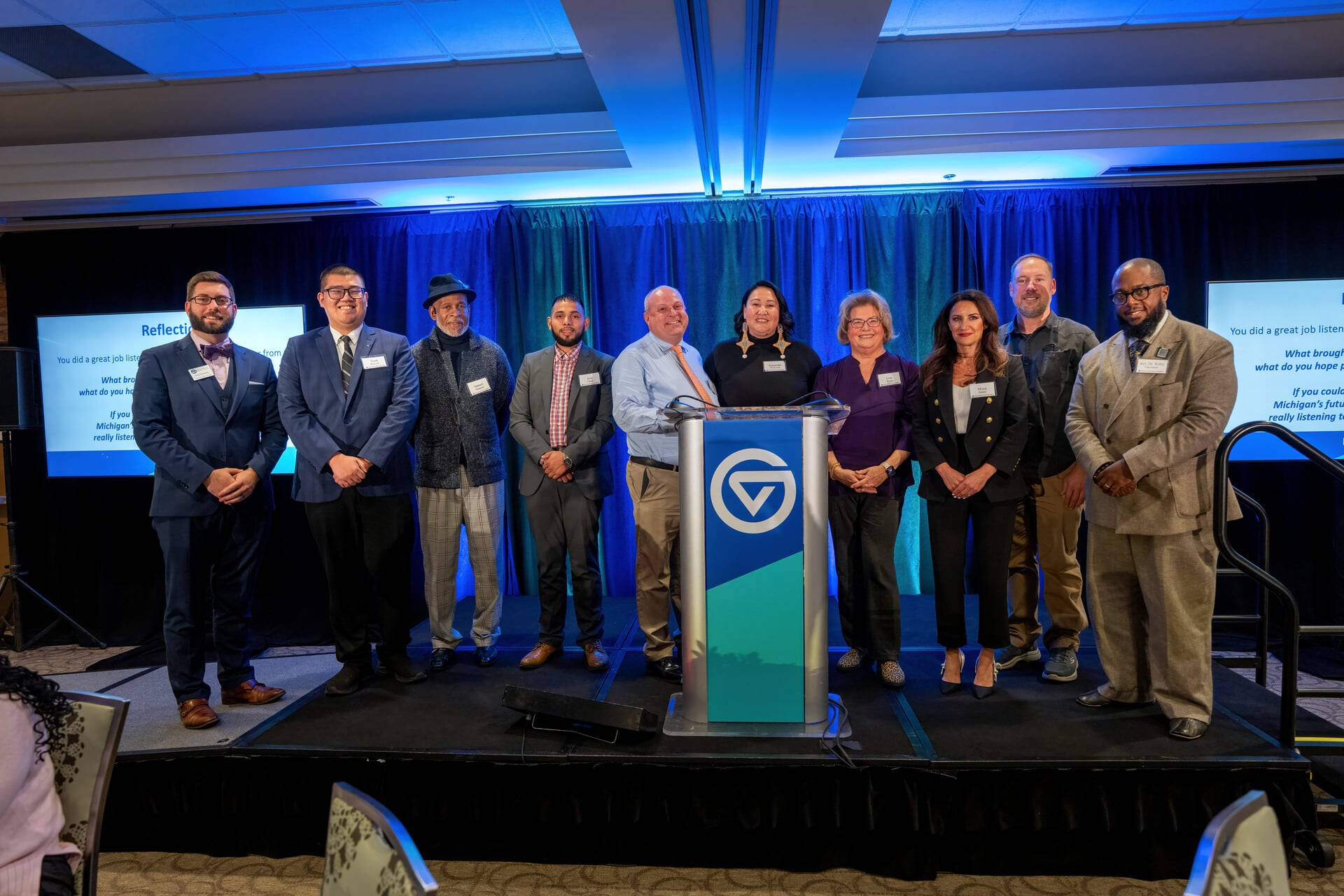 All nine speakers plus Kyle Kooyers, the evening's MC, posing on the Michigan Listens stage.
